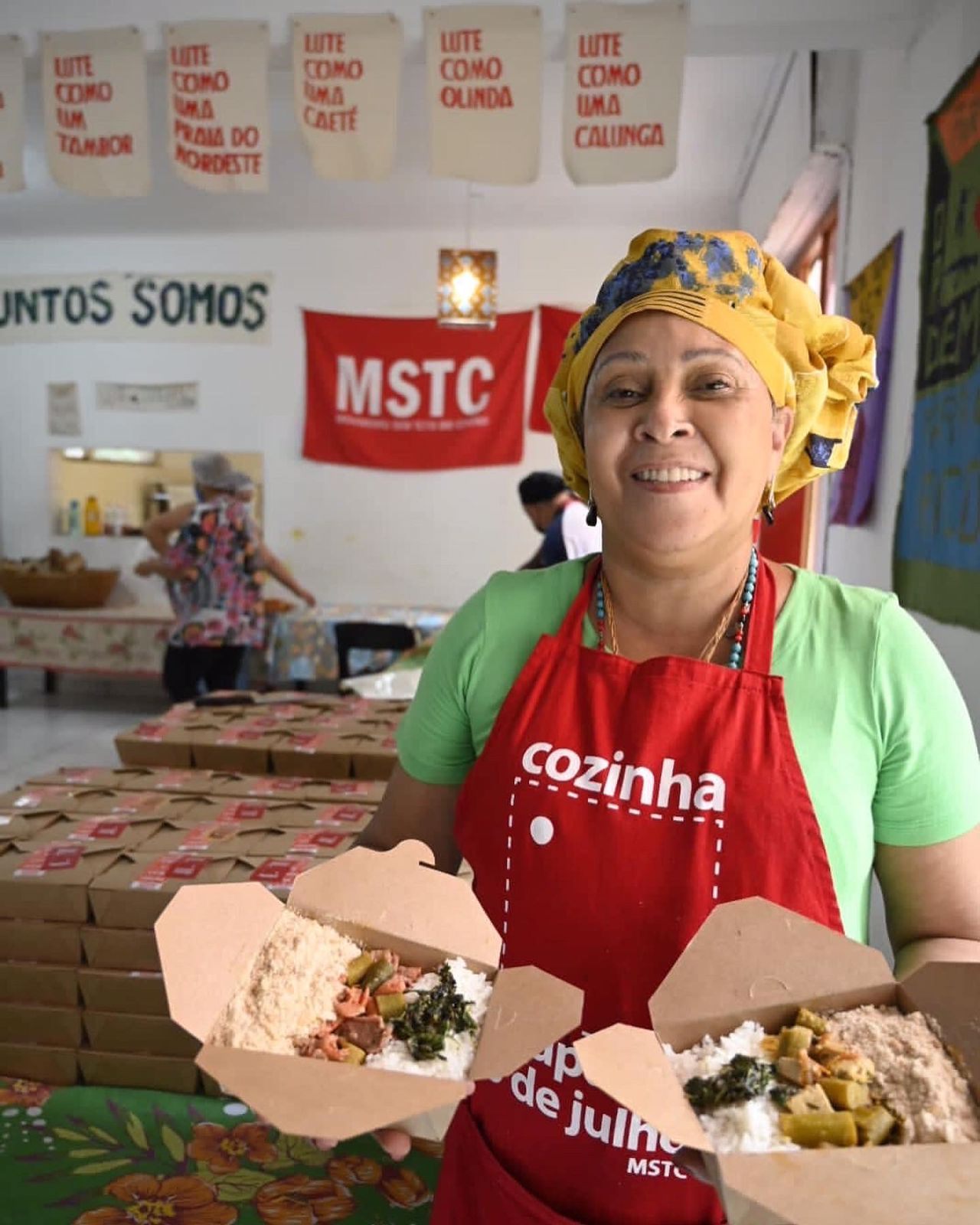 Cozinheira de avental vermelho escrito Cozinha segura duas marmitas montadas; ao fundo, bandeira do MSTC e pilhas de embalagens.