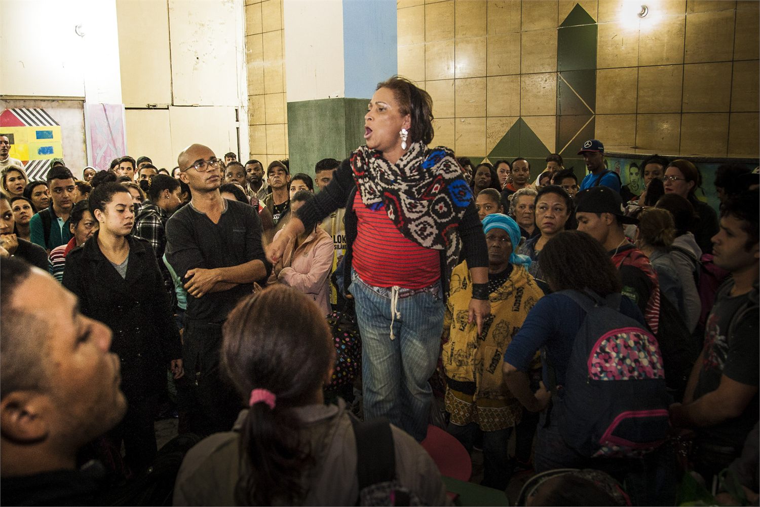 Carmen Silva fala em pé, com o braço estendido, cercada por dezenas de pessoas que a escutam durante uma assembleia.