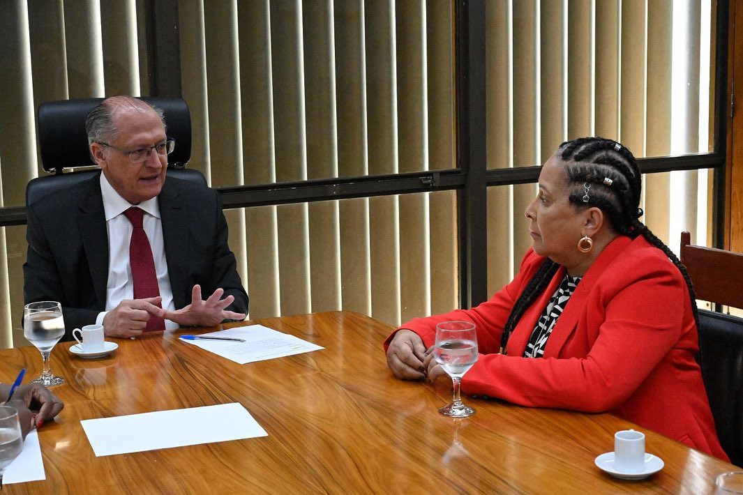 Carmen Silva sentada a uma mesa de reunião, de blazer vermelho, em conversa de trabalho.