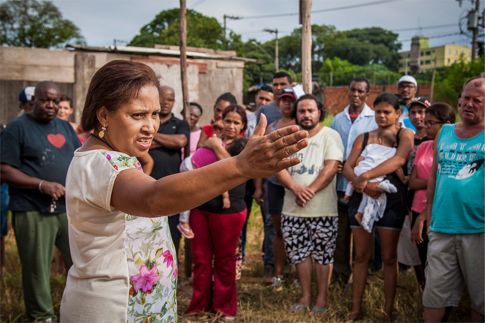 Carmen Silva gesticula ao falar para um grupo de moradores reunidos ao ar livre, num terreno com construções baixas ao fundo.